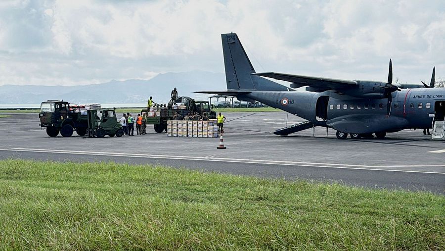 Avion militar francés descargando carga en pista de aterrizaje con ayuda de camiones y personal.  Se observan cajas y bidones.  Posible operación de ayuda humanitaria.
