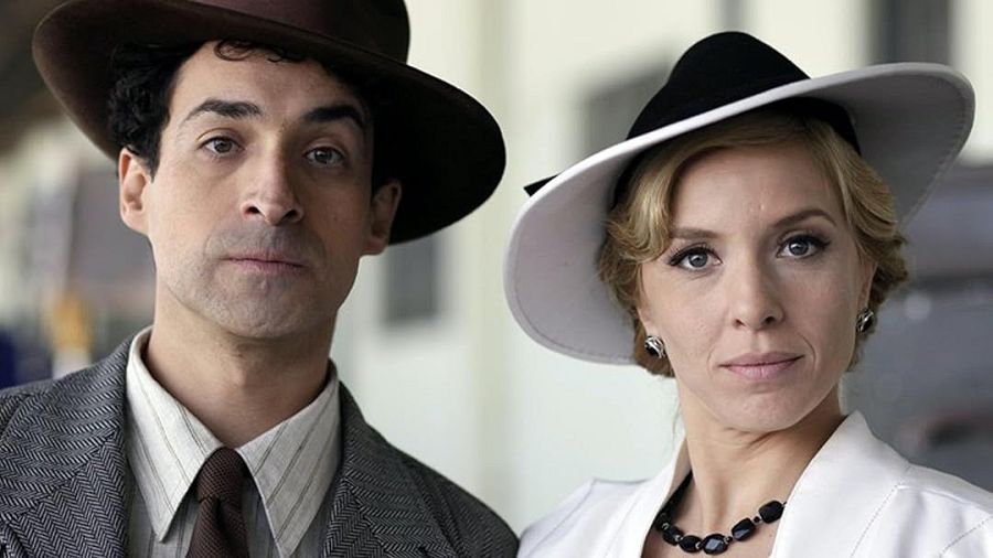 Dos actores, él con traje gris y sombrero, ella con traje blanco y sombrero de ala ancha, miran a cámara con expresión seria.  Ambientación de mediados del siglo XX.