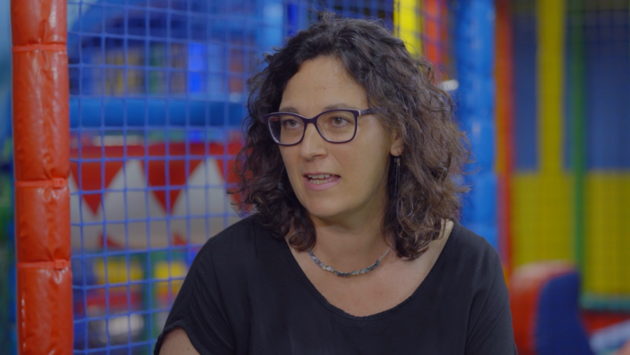 Mujer con gafas y cabello rizado oscuro habla en un parque infantil de colores, posiblemente sobre la tristeza postparto.