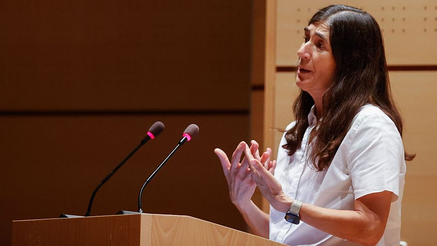 Conferencia impartida por una mujer con camisa blanca y reloj inteligente, de pie tras un atril con dos micrófonos, gesticulando durante su discurso.