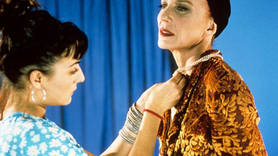 Escena de película con dos actrices: una con blusa azul y flores, coloca un collar a otra con prenda oscura y sombrero.