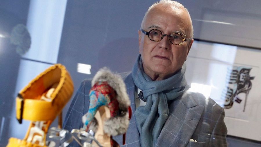 Manolo Blahnik, con gafas redondas y americana a cuadros, posa junto a zapatos de diseño amarillo y azul, y un dibujo de un zapato.