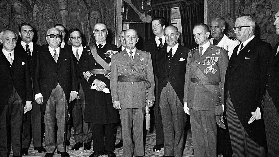 Luis Carrero Blanco, en uniforme militar, posa en una foto con otros hombres en un interior decorado.  Acto oficial durante el franquismo, probablemente anterior a 1973.