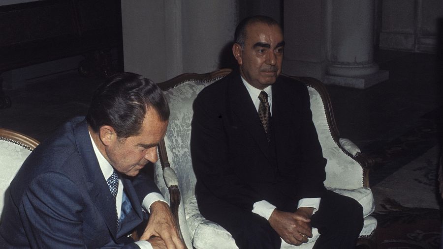 Reunión formal entre dos hombres en trajes. Uno viste traje azul marino y mira hacia abajo; el otro, traje oscuro, mira al frente o a su compañero. Ambiente elegante.