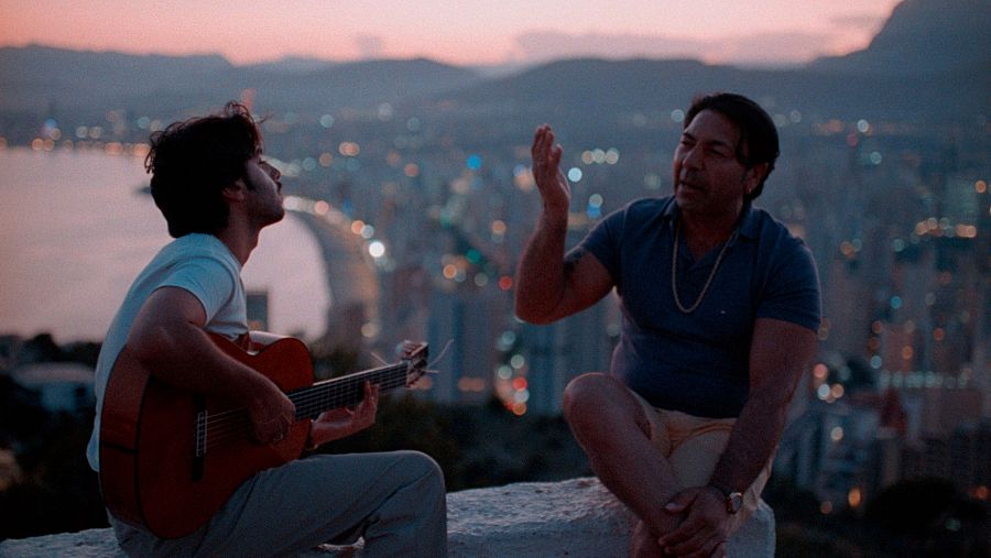 Actuación musical nocturna de un padre y un hijo. Uno toca la guitarra española mientras el otro canta, con un fondo de ciudad costera iluminada.
