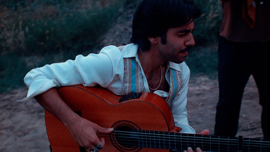 Hombre tocando guitarra española, camisa blanca y chaleco, en exterior.  Concentración en su expresión.  Parte de otra persona visible al fondo.