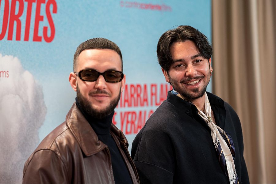 Dos hombres sonríen a la cámara durante la promoción de un documental. Uno viste chaqueta de cuero y gafas; el otro, camisa y pañuelo. El fondo es un cartel azul con texto rojo.