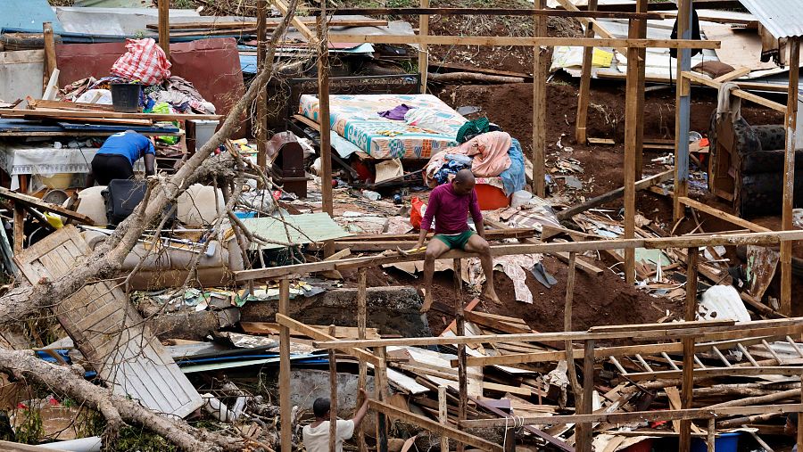 Escena de devastación tras un ciclón: escombros, estructuras dañadas y tres personas, una con camiseta azul, otra roja y otra de color claro, trabajando en la limpieza y reconstrucción.
