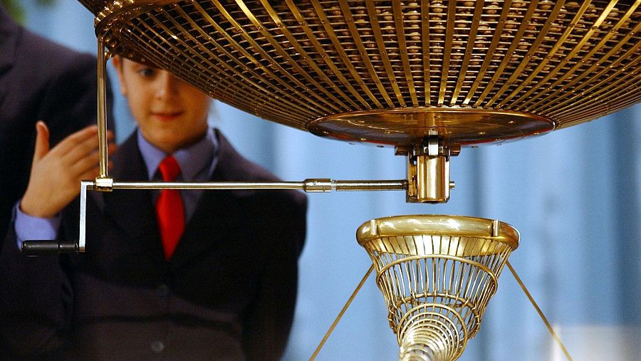 Niño con traje oscuro y corbata roja acciona manivela de un bombón dorado de lotería con rejilla y tubo espiralado. Fondo azul.