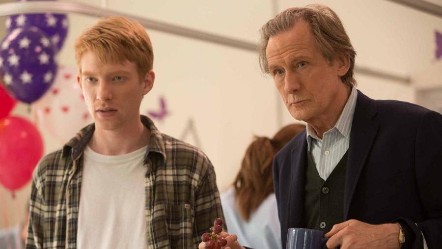 Escena de película con Domhnall Gleeson (camisa a cuadros, uvas) y Bill Nighy (americana, taza). Fondo festivo con globos.