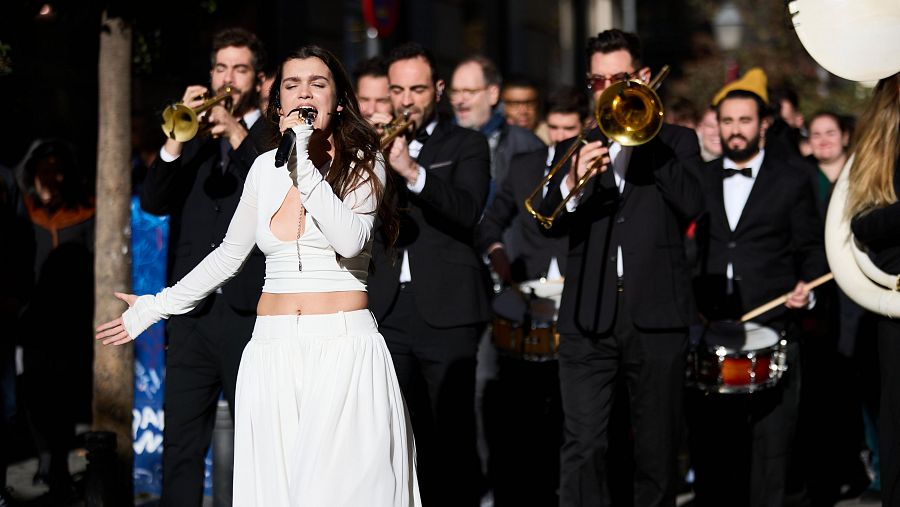 Cantante con atuendo blanco actuando en directo, acompañada de músicos con instrumentos de viento.  Gestos enérgicos mientras canta con un micrófono.