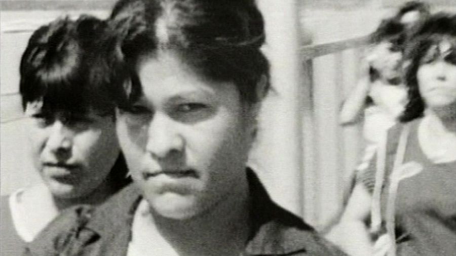 Fotograma en blanco y negro granulado de una obra teatral, mostrando tres mujeres de cabello oscuro en un espacio abierto con una valla al fondo.