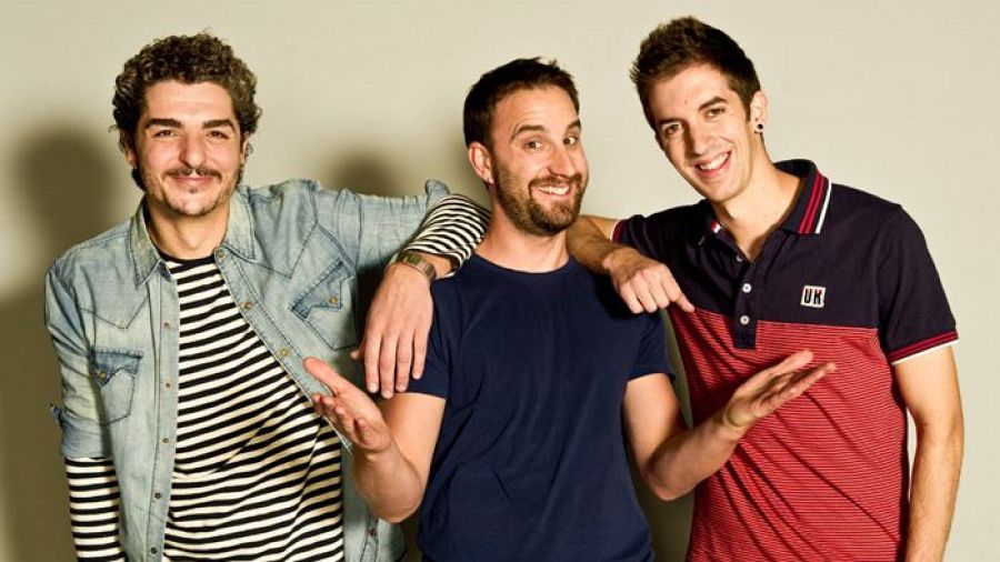 Tres hombres sonríen y posan juntos, mostrando detalles de sus vestimentas: camisa vaquera, camiseta azul marino y polo a rayas. Fondo claro.