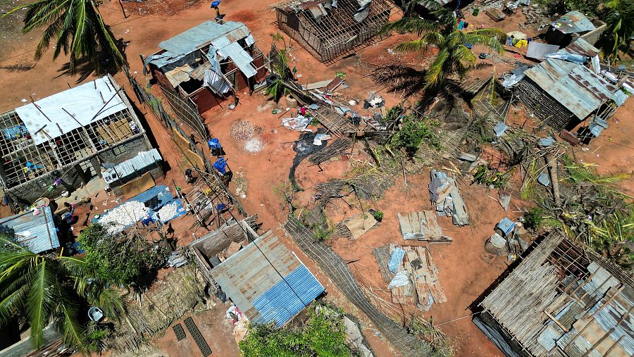 Vista aérea de un poblado afectado por un ciclón: viviendas con techos dañados, escombros y personas entre los restos.  Daños significativos en estructuras de madera y metal.
