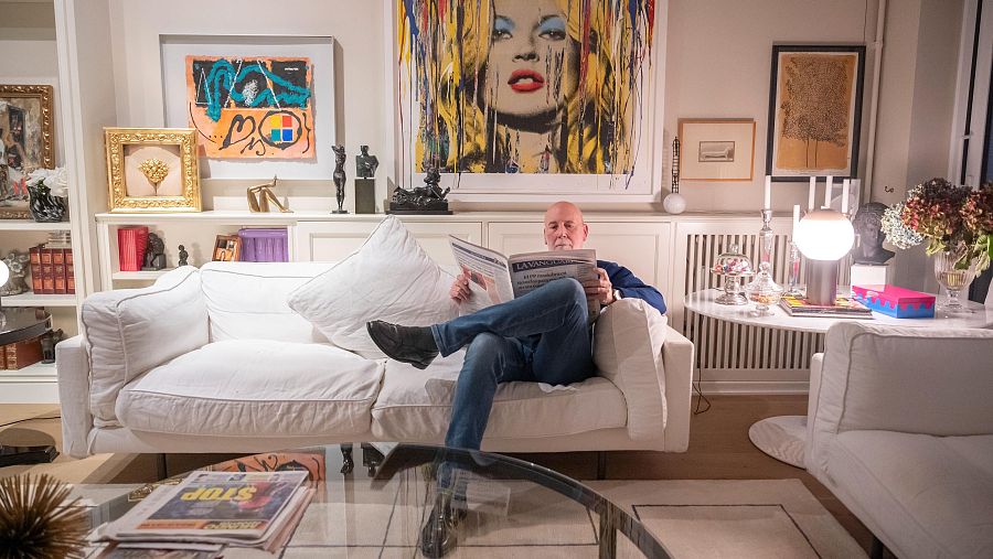 Hombre leyendo La Vanguardia en un sofá blanco, rodeado de libros, cuadros y objetos decorativos.  Ambiente relajado en una sala de estar.
