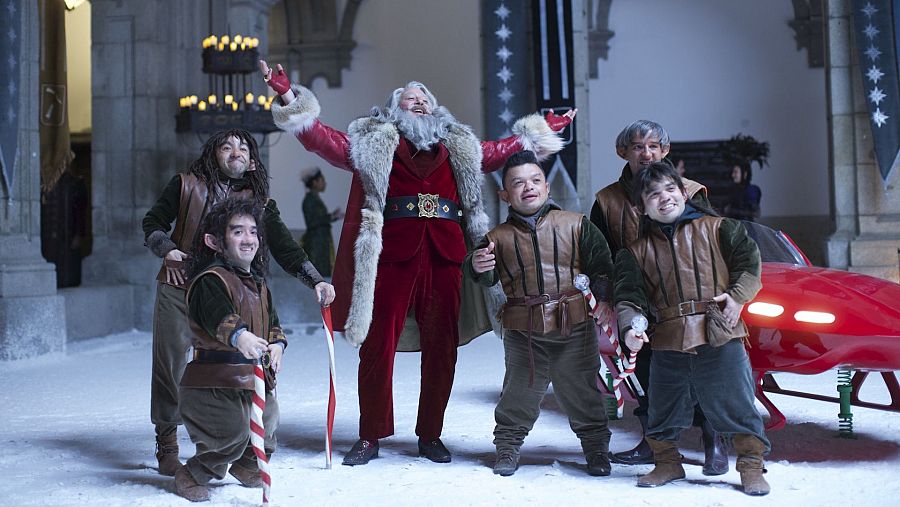 Santa Claus, con traje rojo y barba blanca, rodeado de cuatro personajes medievales con bastones de caramelo, en un patio nevado con un vehículo rojo. Escena de la película 