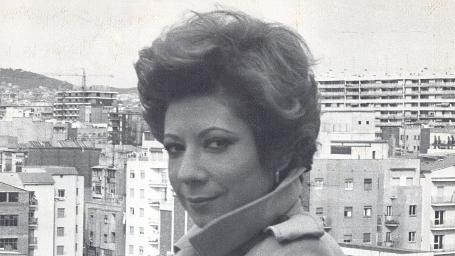 Retrato en blanco y negro de Nuria Feliu; abrigo de cuello alto, pelo corto y rizado, sonrisa leve. Fondo urbano visto desde altura.