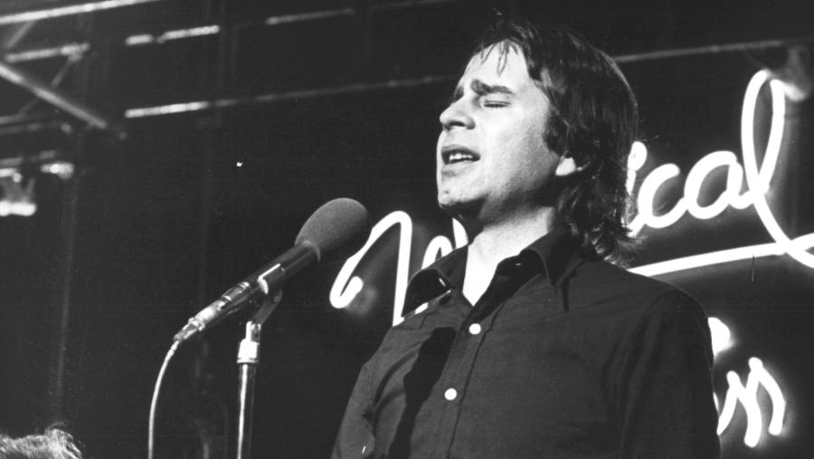 Ovidi Montllor, en blanco y negro, canta al micrófono con los ojos cerrados, vistiendo camisa oscura. Actuación en escenario con rótulo luminoso de fondo.