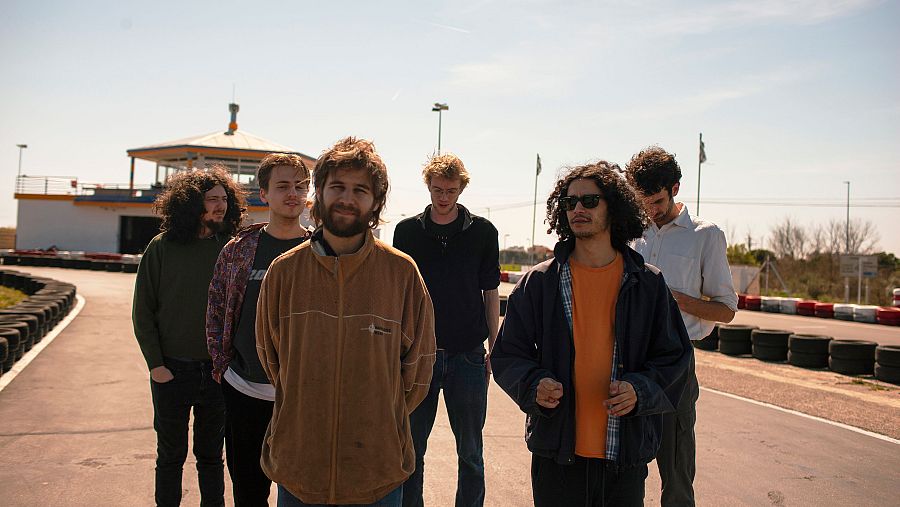Jóvenes en pista de karts, con estructura circular blanca al fondo y neumáticos apilados. Día soleado. Posible videoclip o sesión fotográfica.