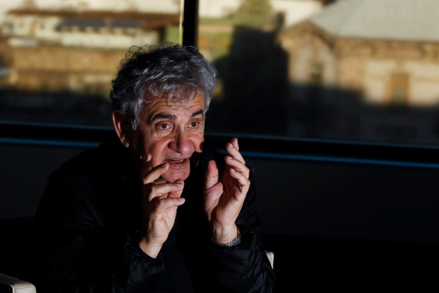 Fotografía de Bernardo Atxaga sentado, con chaqueta oscura y cabello canoso, gesticulando mientras parece hablar. Fondo borroso con indicios de una ventana.