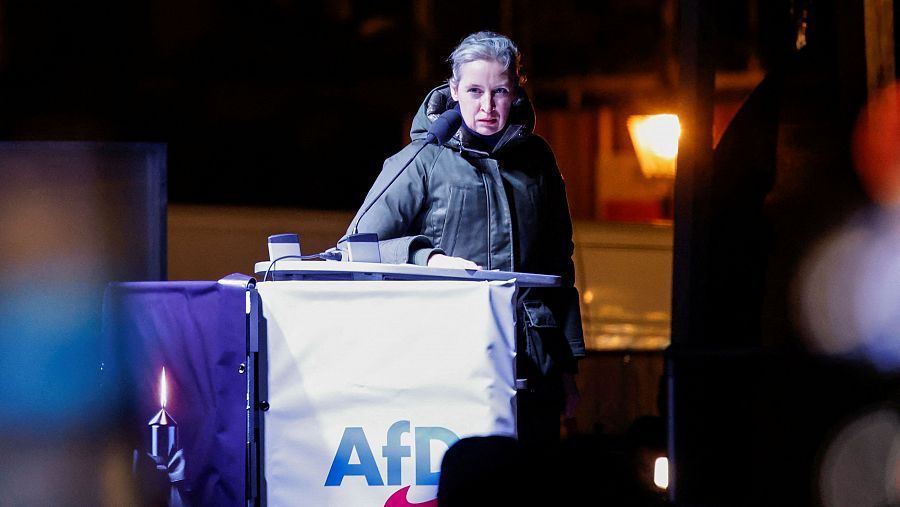 Mujer con abrigo oscuro ofreciendo un discurso tras un atril con el logo de AfD; vela encendida visible; ambiente nocturno.