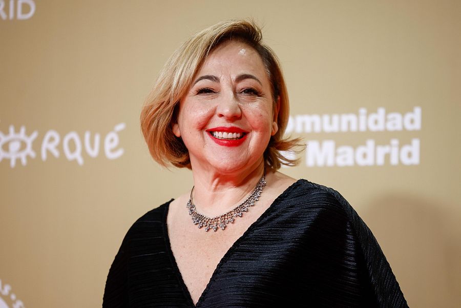 Carmen Machi, en los Premios Forqué, luce blusa negra y collar plateado, sonriendo a cámara. Fondo dorado con logo de los premios y 'Comunidad de Madrid'.