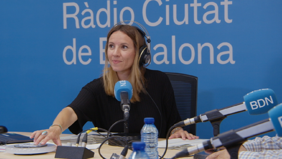 Locutora con auriculares presenta un programa de radio, sentada ante una mesa de mezclas y micrófono.  Sobre la mesa hay papeles y agua.