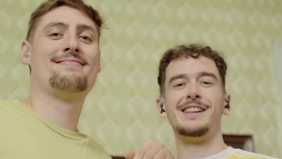 Dos jóvenes, con camisetas de colores distintos y pelo corto, sonríen a cámara.  Ambiente interior.