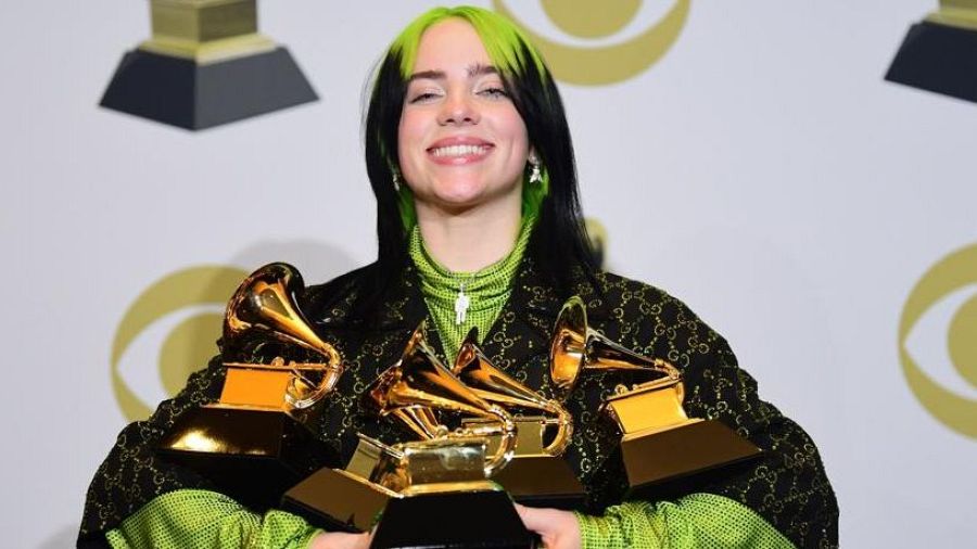 Billie Eilish sonríe sosteniendo múltiples premios Grammy en los Grammy 2020.  Lleva un conjunto Gucci y cabello verde y negro.