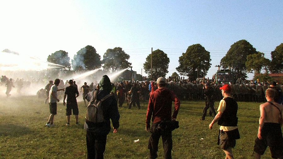 Fotografía de una protesta: manifestantes dispersos, enfrentamiento con policía antidisturbios usando cañones de agua, dos observadores (uno con cámara).