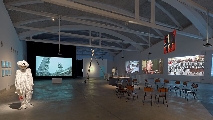 Exposición con oso polar, proyecciones de industria y protestas, instalación triangular y sillas.  Ambientación industrial, temática medioambiental.