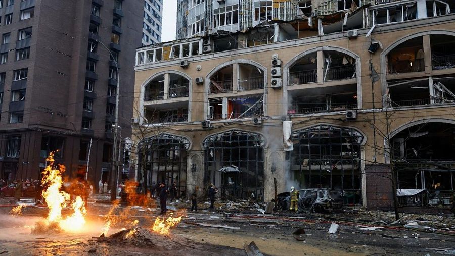 Edificio dañado por fuego, escombros en la calle y vehículos destruidos.  Personas, incluyendo posibles bomberos, presentes en la escena.