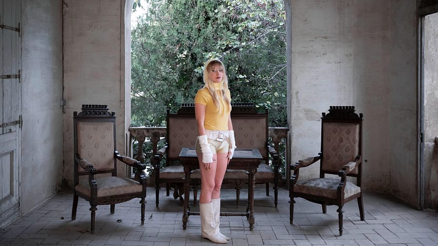 Maria Hein, con jersey amarillo y pantalones cortos, posa en un interior antiguo con sillones y una ventana con vista a un jardín.