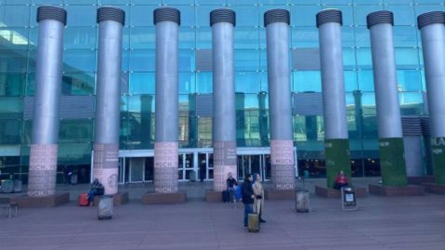 Zona de fumadores con columnas metálicas, bancos, maletas y ceniceros. Personas sentadas y de pie. Cartel ilegible en una columna.