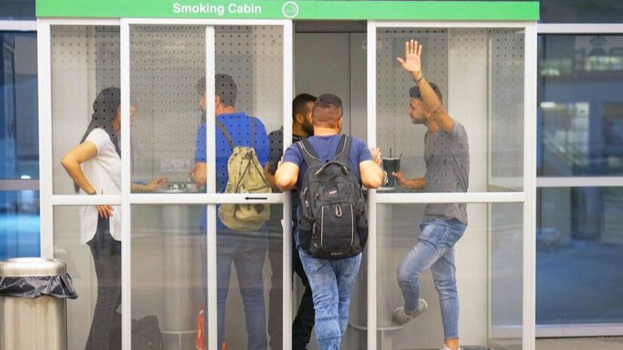 Personas fumando en una cabina de aeropuerto con estructura metálica y paneles de vidrio.  Se observa una papelera cercana.
