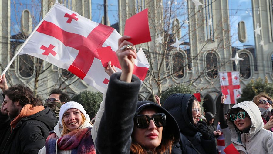 Manifestantes en Georgia expresan su descontento con tarjetas rojas y banderas nacionales frente a un edificio.  La protesta parece dirigida a una figura de autoridad.