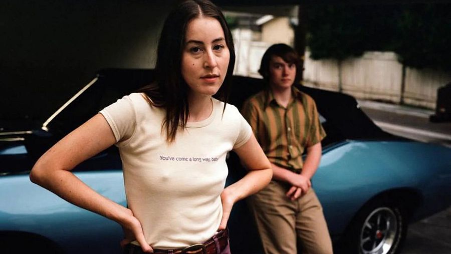 Alana Haim en escena de película, camiseta beige con eslogan, manos en caderas, junto a joven, apoyados en coche descapotable. Estética años 70.