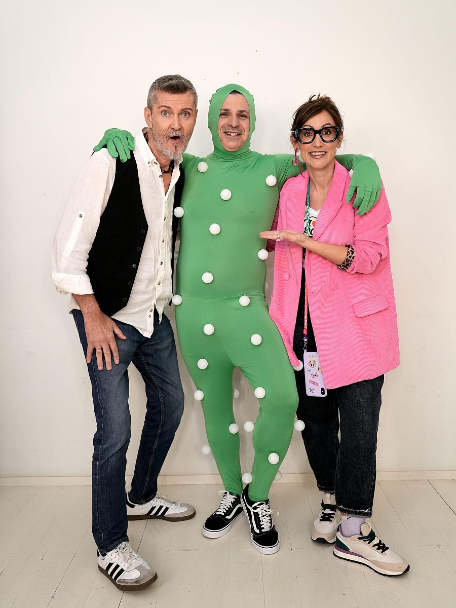 Tres presentadores posan para una foto promocional.  Uno viste camisa blanca y chaleco, otro un traje verde con bolas blancas, y la tercera una chaqueta rosa.