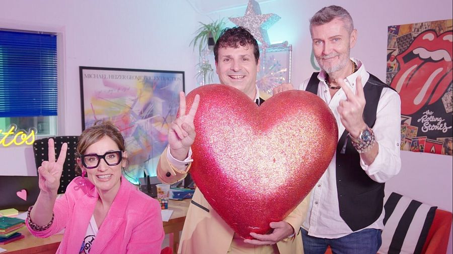 Presentadores de 'Cachitos' haciendo el símbolo de la paz con un gran corazón rojo en un plató decorado con motivos de los 90.