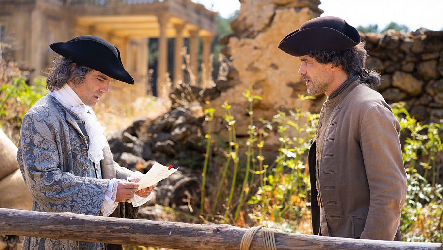 Dos hombres en trajes de época, uno leyendo una carta con expresión seria, en un entorno rural con ruinas y una gran construcción al fondo.  Escena soleada.