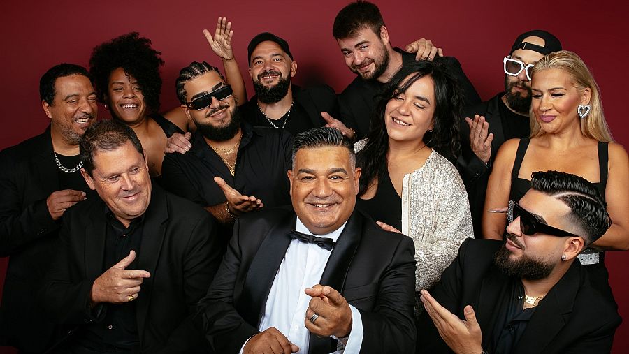 Grupo de once personas sonrientes posando para una foto con fondo granate. Un hombre en traje negro y pajarita destaca en el centro, señalando a la cámara.