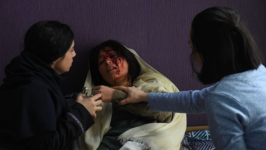 Tres mujeres, una herida y dos que la asisten, en una escena dramática de interior con pared morada.  Se aprecian manchas de sangre y una tela estampada.