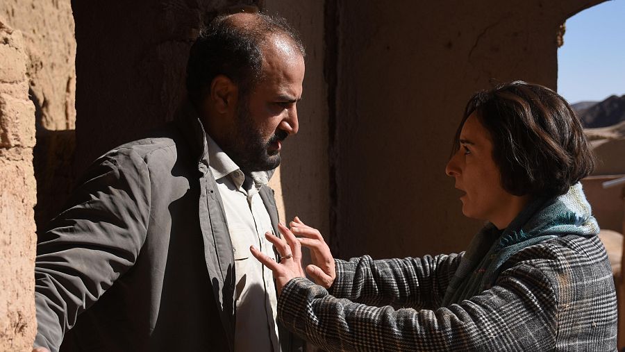 Escena de película: hombre y mujer con expresiones serias interactúan en un espacio de adobe iluminado por una abertura que muestra un paisaje árido.