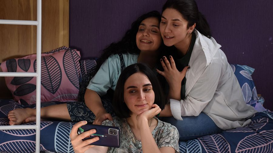 Selfie de tres mujeres jóvenes en una cama con sábanas estampadas, sonriendo y abrazándose. Fondo morado.