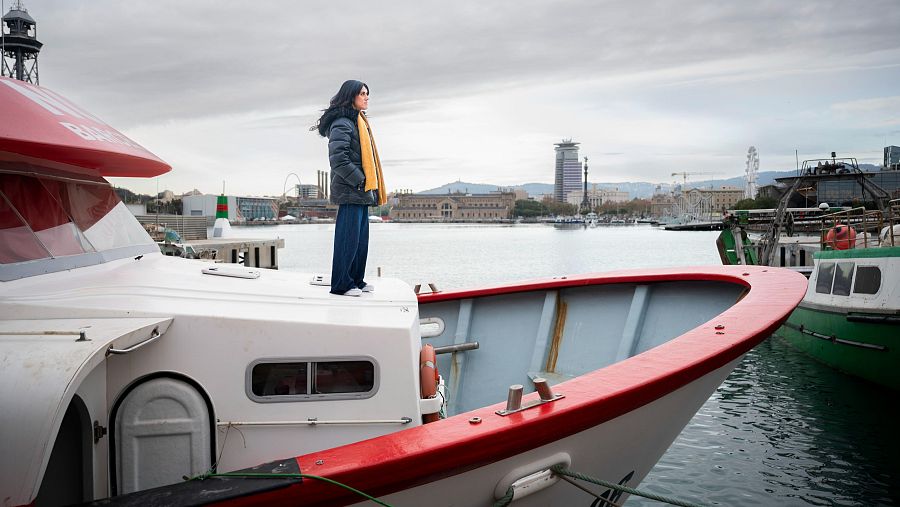 Mujer con abrigo oscuro y pantalones azules en un barco pesquero blanco y rojo en un puerto.  Mira hacia la ciudad, con edificios visibles al fondo.