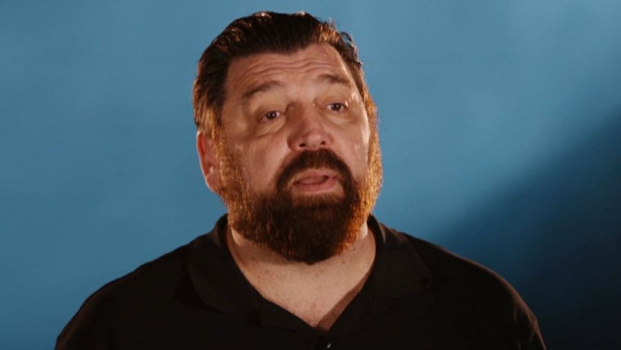 Primer plano de Hovik Keuchkerian con expresión seria, camisa oscura de manga corta y fondo azul.  Parece una entrevista.