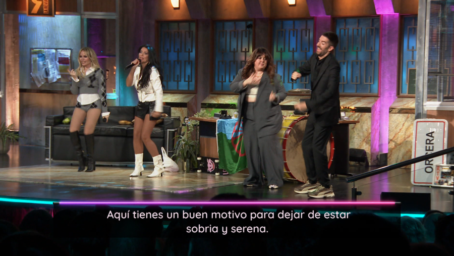 Escena con cuatro personas: dos mujeres, un hombre y una humorista, en un escenario con instrumentos musicales y un letrero de 