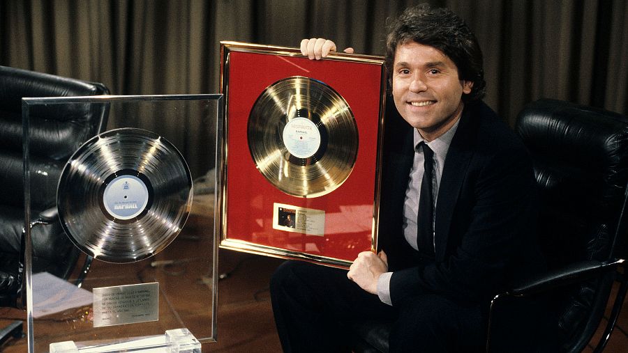 Cantante con traje oscuro sostiene un disco de oro enmarcado y otro plateado, sonriendo a la cámara. Fondo con cortina.