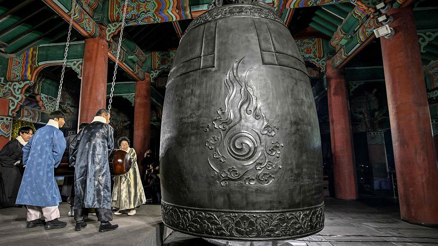 Campana oscura con grabados, personas en hanbok a punto de golpearla en una ceremonia tradicional.