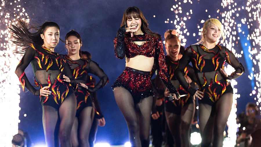 Lisa de BLACKPINK canta con entusiasmo en un escenario iluminado, luciendo un conjunto rojo brillante y rodeada de bailarinas con bodies negros con llamas rojas y amarillas.